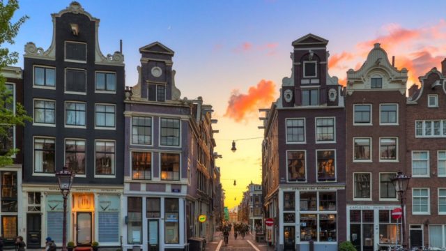Der Sonnenuntergang über den Grachten von Amsterdam bietet einen atemberaubenden Anblick mit einem farbenfrohen Himmel und den ikonischen Gebäuden der Stadt als Silhouette.