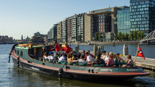 Touristen genießen eine Grachtenfahrt auf einem Boot im modernen Amsterdam, mit Gebäuden am Wasser im Hintergrund.