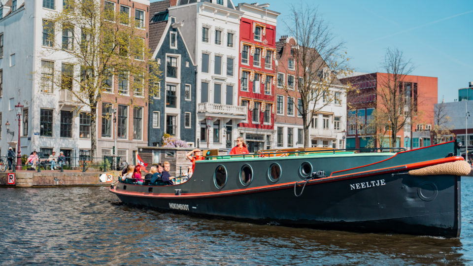 Rondvaartboot op een Amsterdamse gracht met kleurrijke historische huizen op de achtergrond.